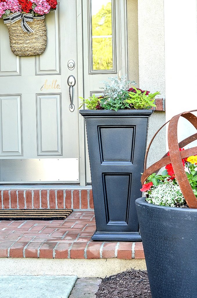 SMALL FRONT PORCH WITH BLACK PLANTERS. LOTS OF FLOWERS IN THE PLANTERS.